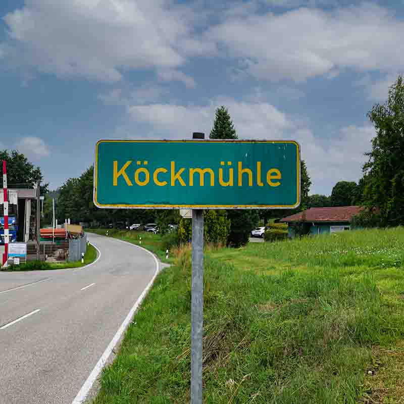 Ein grünes Ortseingangsschild mit der gelben Aufschrift Köckmühle steht mittig im Vordergrund, davor eine leicht kurvige Landstraße.